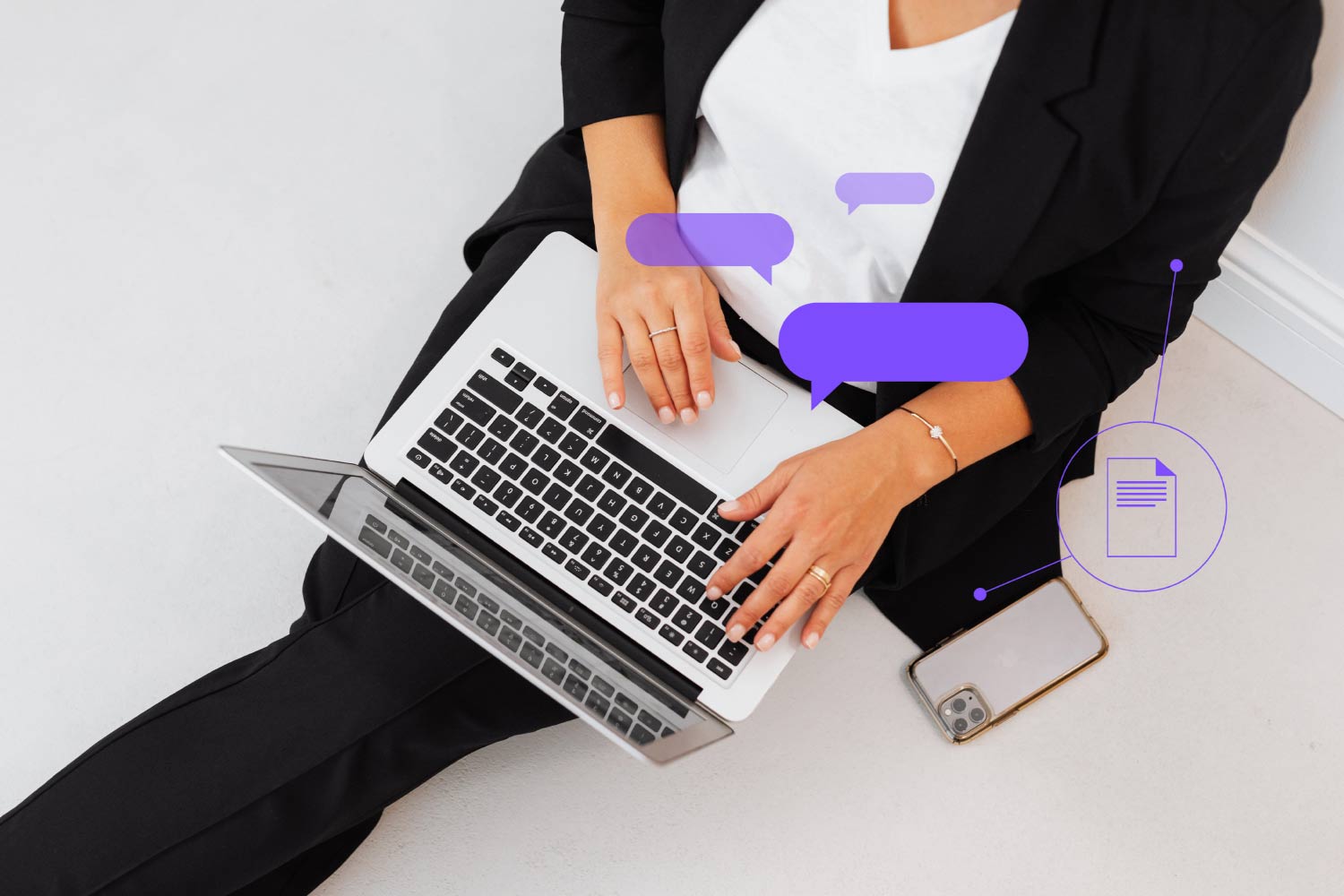 Kobieta siedząca na ziemi, oparta o ścianę, elegancko ubrana. Na kolanach trzyma włączonego laptopa, obok niej na ziemi leży telefon komórkowy