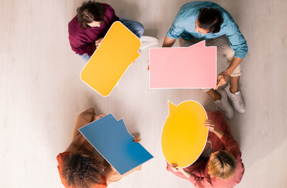 grupa osób siedząca w kole, trzymają plansze w różnych kolorach w kształcie chmurek dialogowych