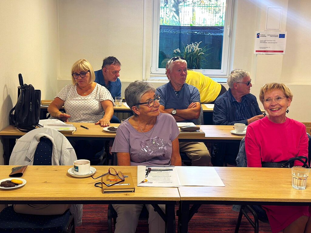 Grupa dorosłych osób siedzi w sali szkoleniowej przy stołach, na których znajdują się notatki, długopisy oraz filiżanki z kawą i talerzyki z ciastkami. Osoby są zaangażowane i skupione na zajęciach, w tle widoczne są materiały informacyjne i okno.